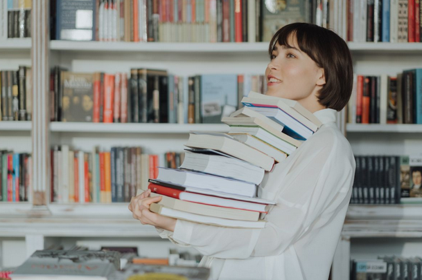 woman standing in front of a book shelf holding stack of books