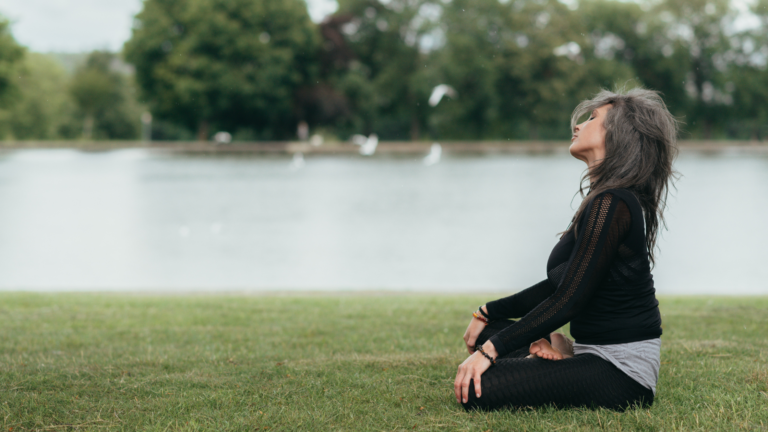A woman sits cross-legged in the grass in front of a lake, meditating.