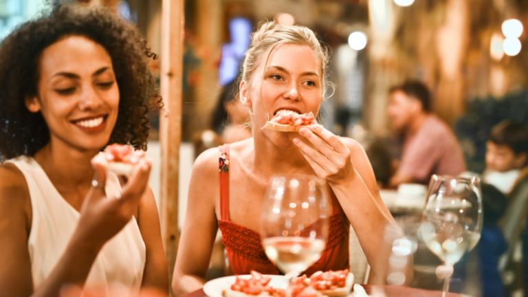 A Black woman (left) and a white woman (center) smile as they sit in a restaurant eating a pizza.