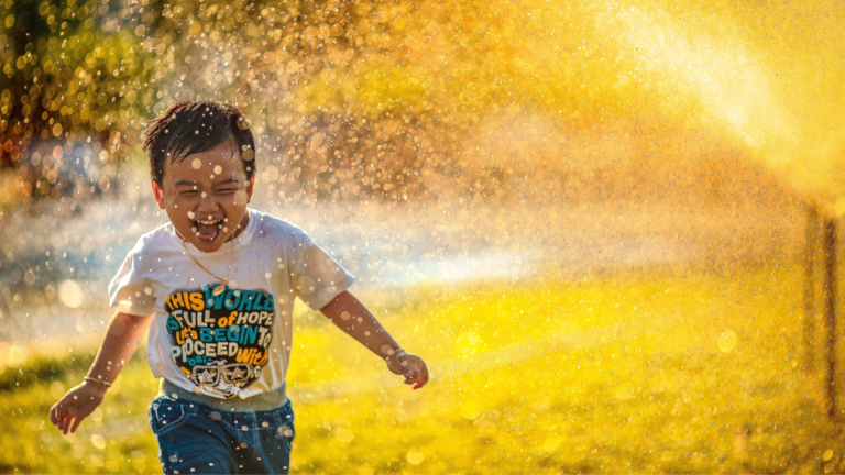 A preschool-aged boy runs through the spray of a sprinkler.